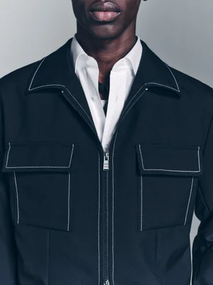 Close-up of a Black male model wearing a dark zip-up jacket with contrast white topstitching over a white collared shirt.