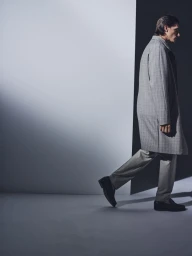 Model Edoardo Sebastianelli walks in profile wearing a grey plaid coat and tailored trousers against a minimalist studio wall.