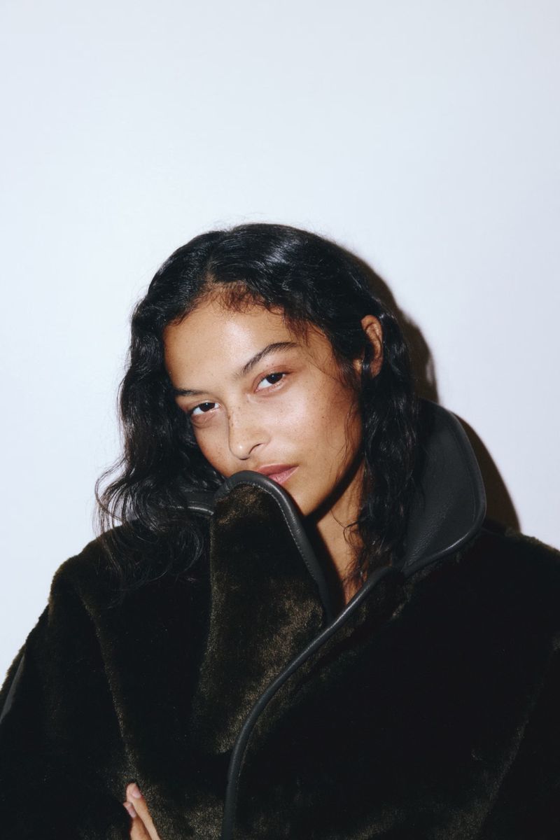 Portrait of a woman with long dark hair wearing a brown fur coat against a white background in a fashion or beauty context.