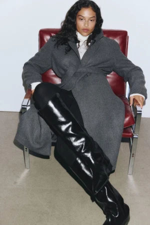 Model Devyn Garcia seated on a red leather armchair wearing a grey coat and black boots for H&M AW2025