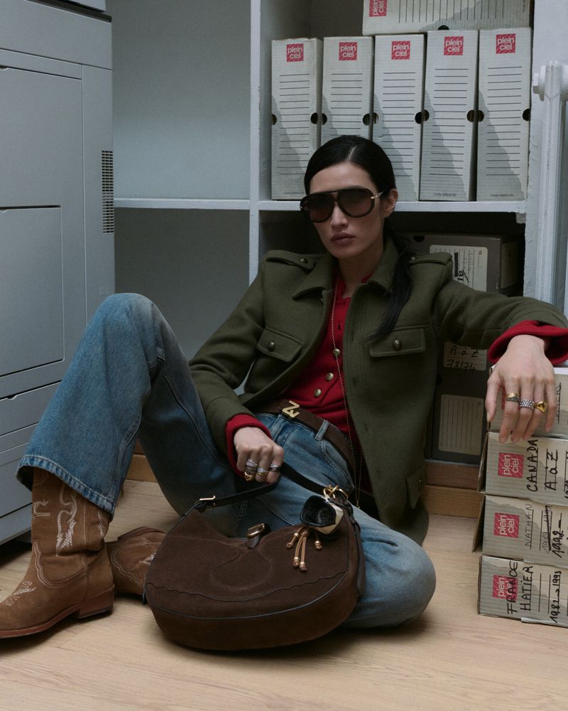Stylish woman poses indoors wearing an olive-green jacket, red top, blue jeans, and brown cowboy boots, holding a brown purse.