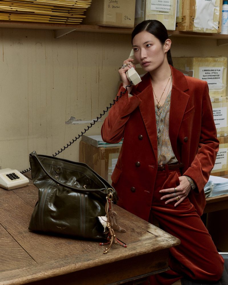 A woman in a rust-colored suit holds a vintage telephone with a purse on a wooden table in an indoor office setting.