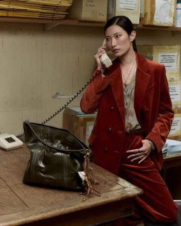 A woman in a rust-colored suit holds a vintage telephone with a purse on a wooden table in an indoor office setting.