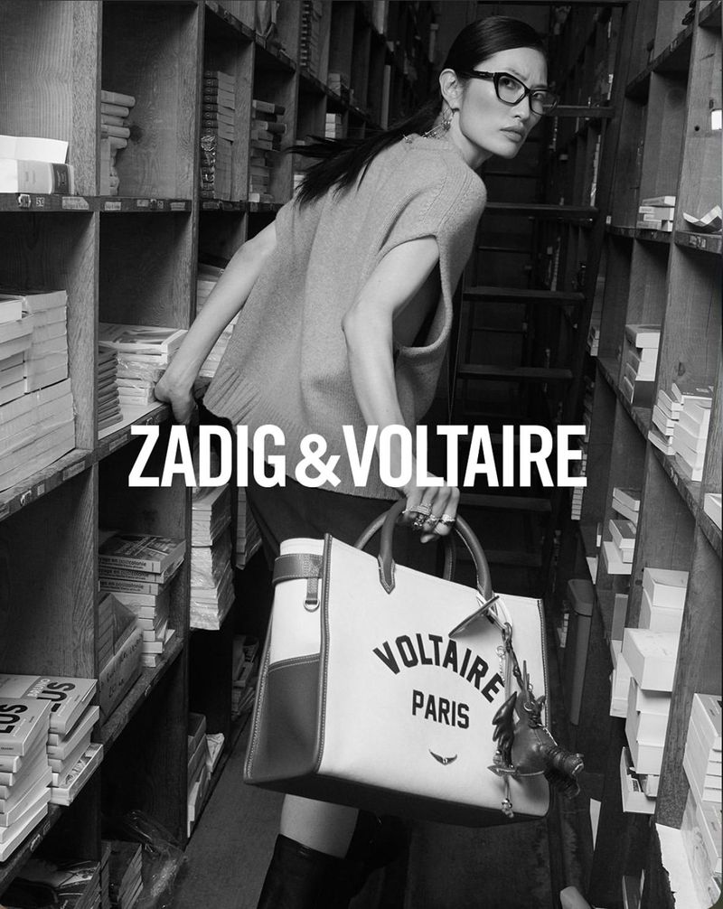 Fashion advertisement featuring a woman posing indoors with a purse, wearing glasses and a sweater, with bookshelves in the background.