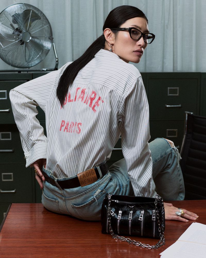 Stylish woman in glasses sits on desk with black handbag. Striped shirt with text, light blue jeans, and office fan and file cabinet.