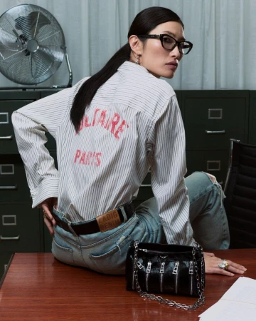 Stylish woman in glasses sits on desk with black handbag. Striped shirt with text, light blue jeans, and office fan and file cabinet.