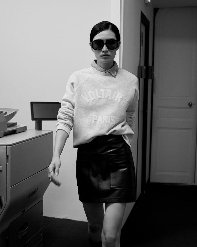 A slender woman with sunglasses in a grey sweatshirt and black mini skirt stands indoors near a printer in a monochrome image.