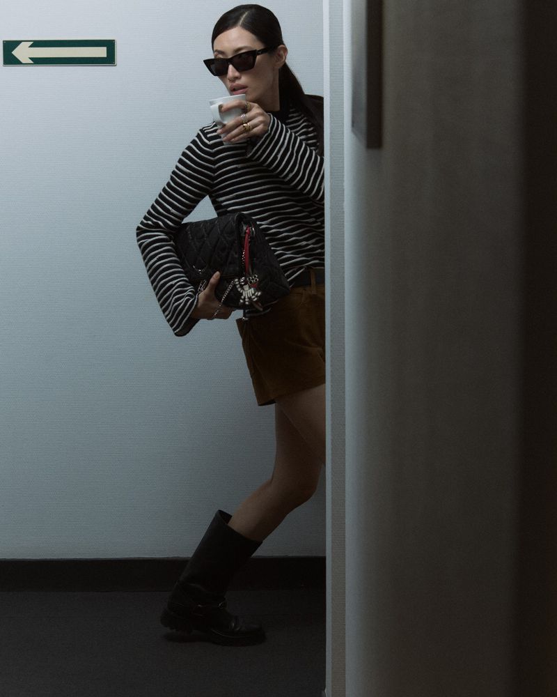Stylish woman in black and white top, brown shorts, and boots holding a cup and purse, posing indoors against a wall.