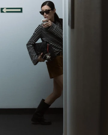 Stylish woman in black and white top, brown shorts, and boots holding a cup and purse, posing indoors against a wall.