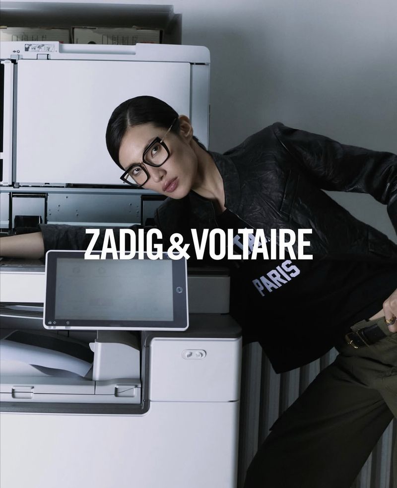An Asian woman with glasses stands next to an office copier with a blank tablet, wearing a black tee and jacket in a fashion editorial.