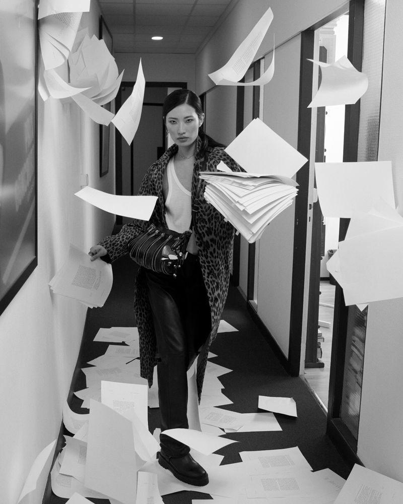 Woman in leopard print coat in an office hallway with papers falling and on the floor in a black and white editorial fashion shot.