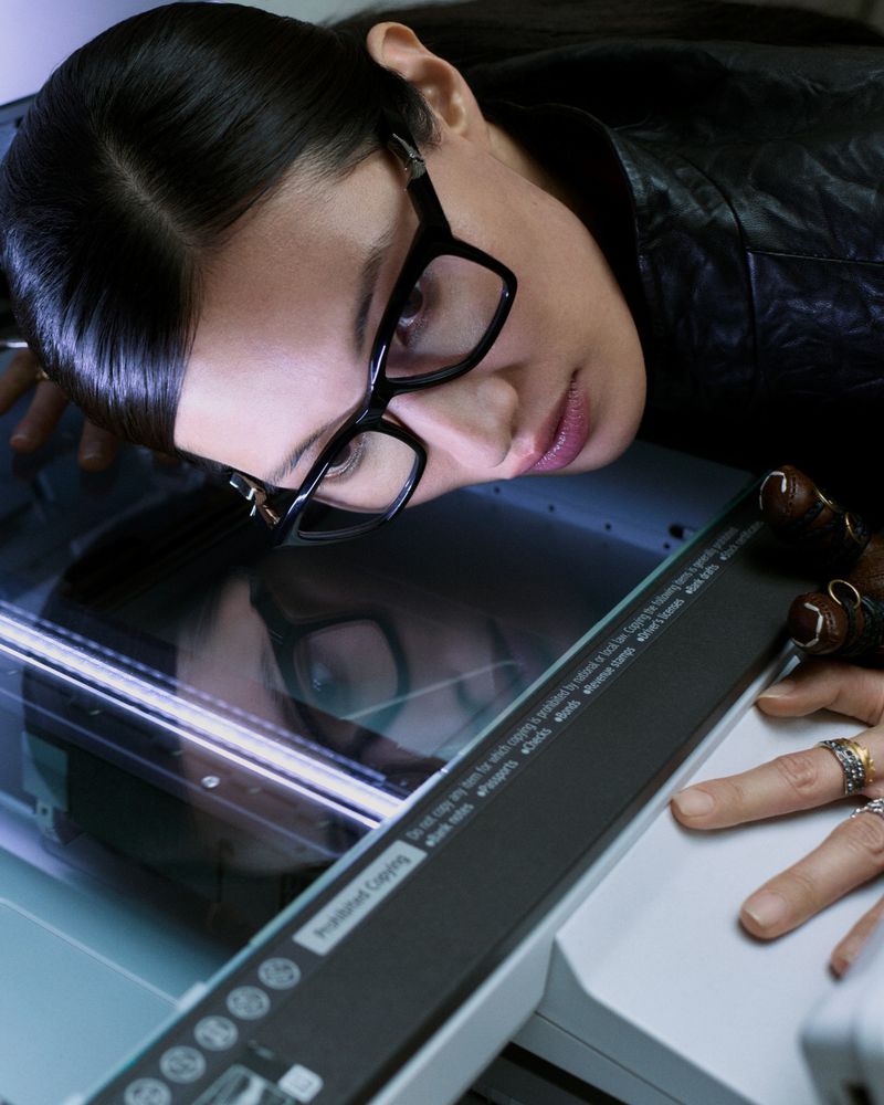 Woman with glasses leans on a printer, her face reflected in the glass, wearing a black textured jacket, showing rings on her fingers.