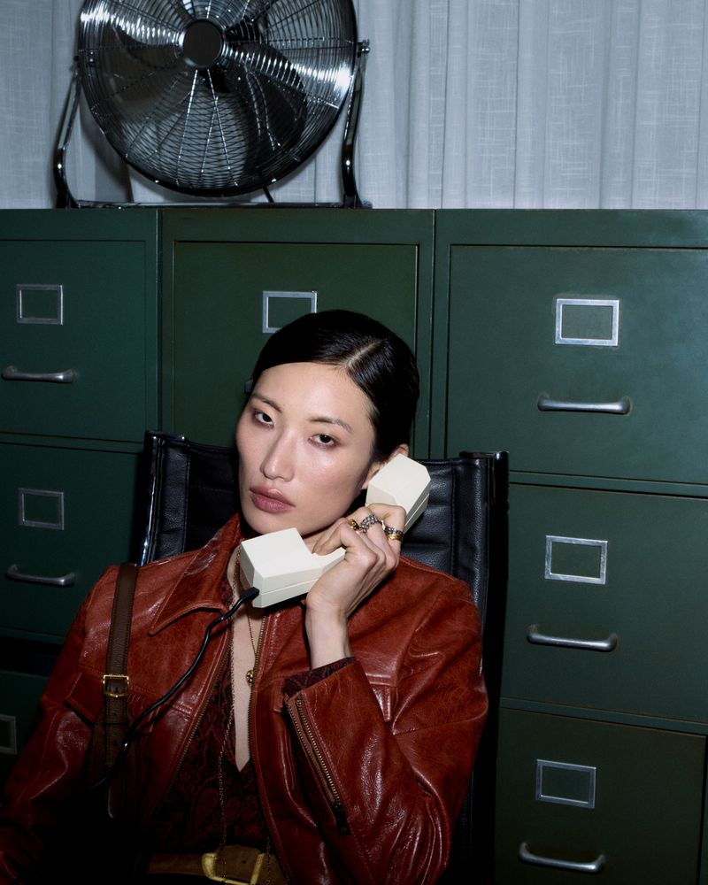 Asian woman in a red leather jacket sits on a chair while holding a vintage telephone with a filing cabinet and fan in the background.