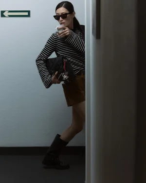 Model Nuri Son in sunglasses, a striped long-sleeve top, brown shorts, and black boots holds a coffee cup and bag in a hallway.