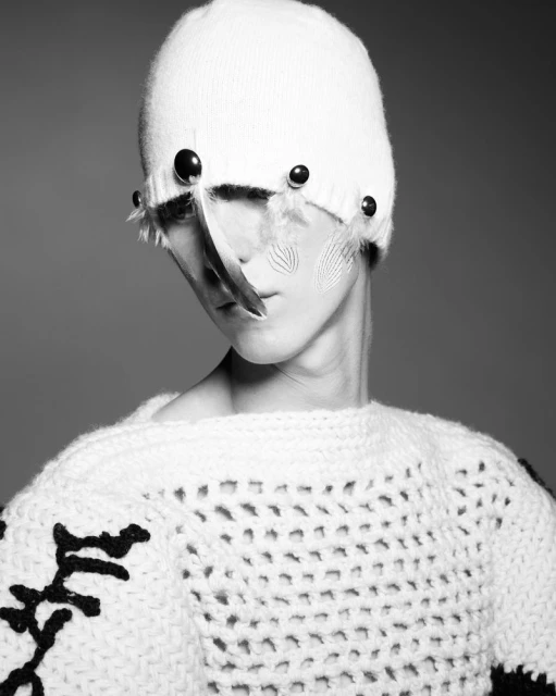 Black-and-white portrait of a person wearing a white knit hat and sweater with black accents, styled with feathers.