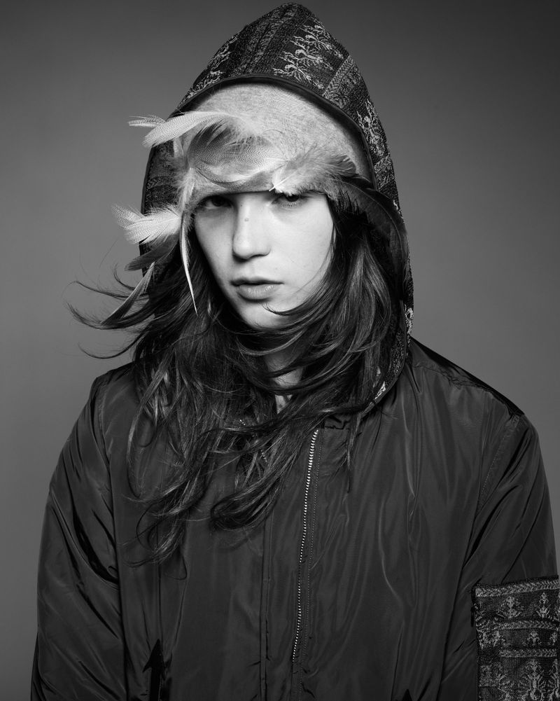 Monochrome portrait of a young adult with long hair wearing a hooded jacket and feather headdress against a solid grey background.