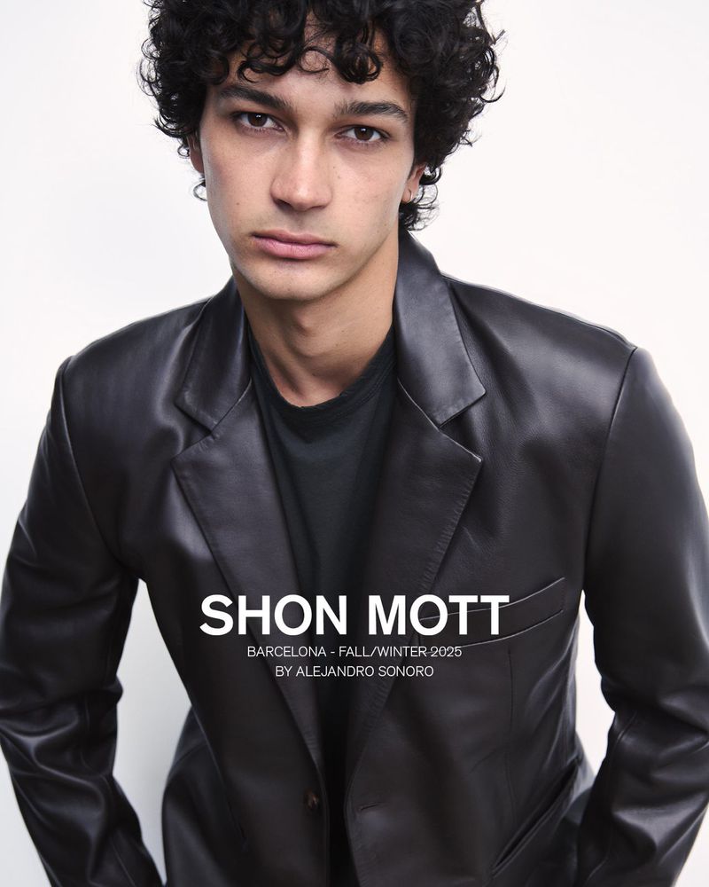 Close-up studio portrait of a young adult male with curly hair wearing a dark jacket, showcasing a fashion editorial style and beauty focus.