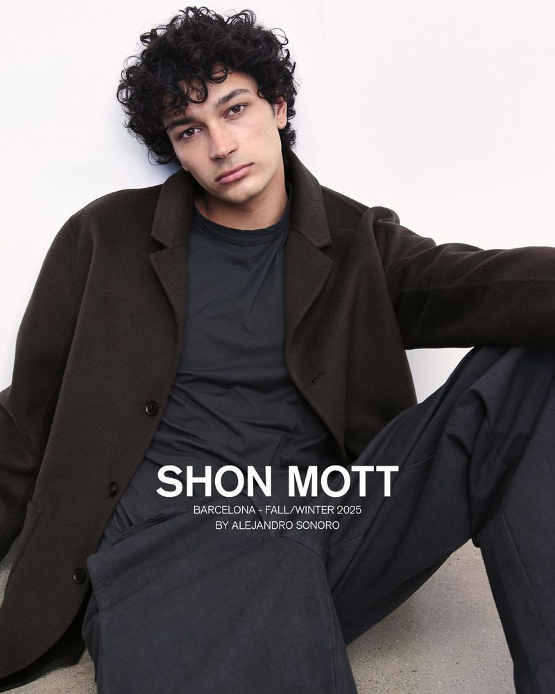 A young male model with dark curly hair wearing a dark brown coat and black t-shirt seated against a plain white background.