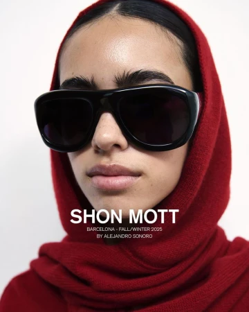 Close-up studio shot of a young adult female model wearing oversized black sunglasses and a textured red hood, set against a plain background.