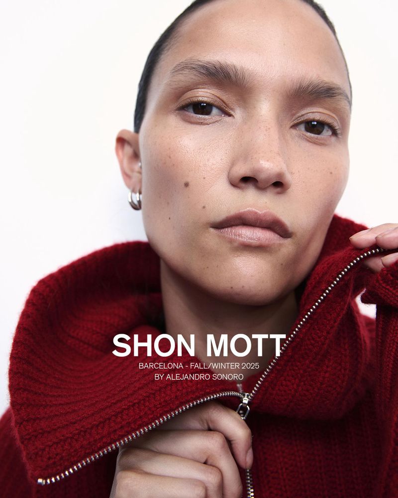 Close-up portrait of a woman with dark hair wearing a red sweater with a silver zipper, against a plain white background. Fashion advertisement.