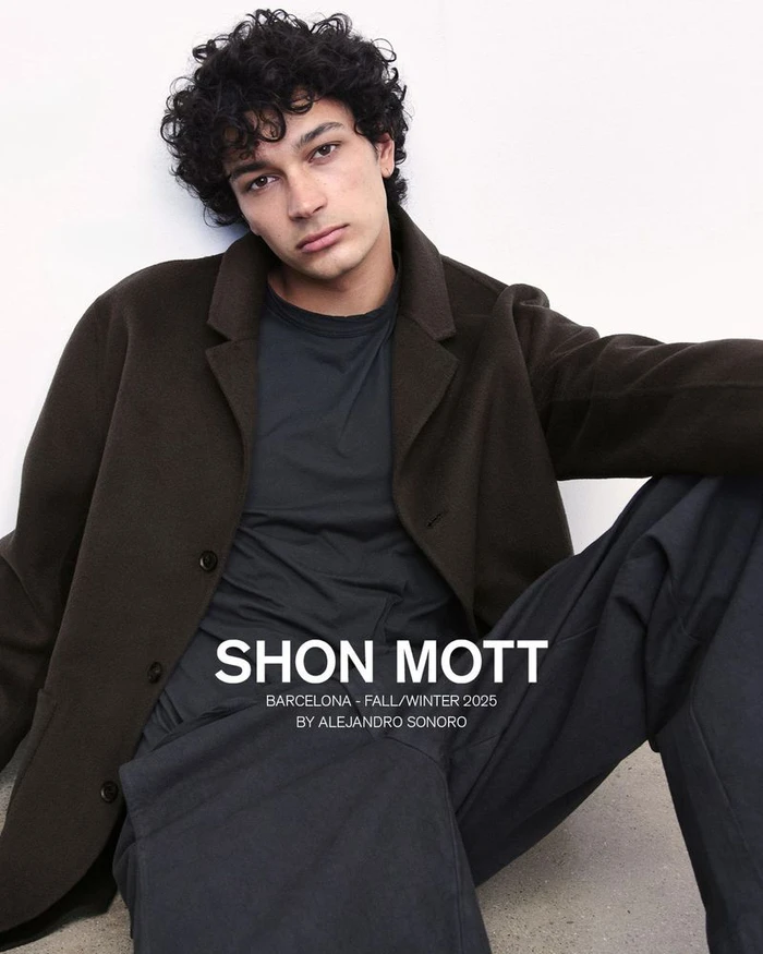 A young male model with dark curly hair wearing a dark brown coat and black t-shirt seated against a plain white background.