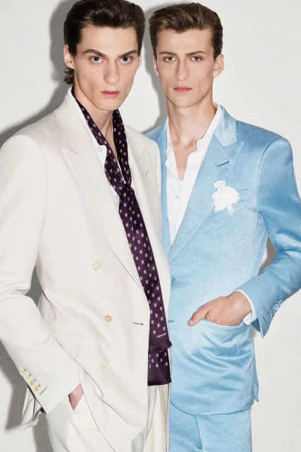 Two men in formal suits, one in light blue and the other in cream, stand indoors against a white background, showcasing fashion apparel.