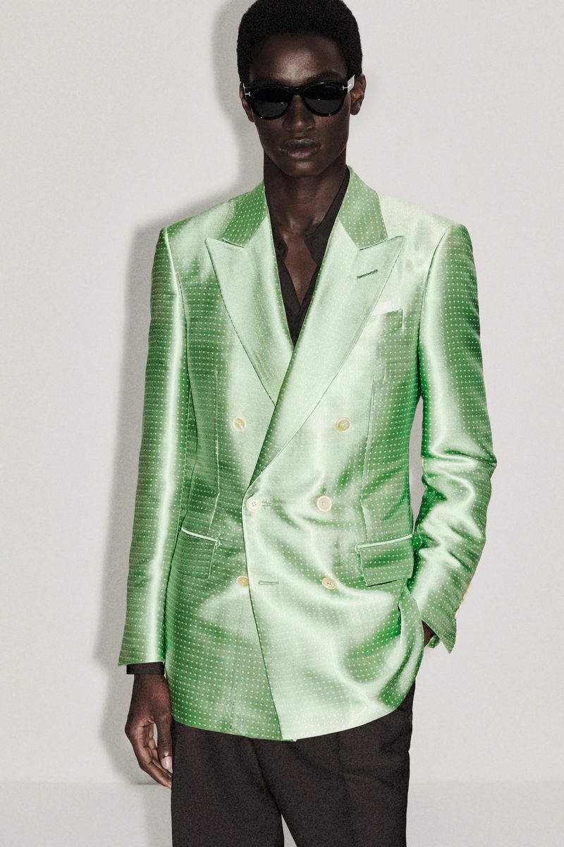 Adult male model wearing a green satin double-breasted blazer and dark pants in a fashion shot against a white background.