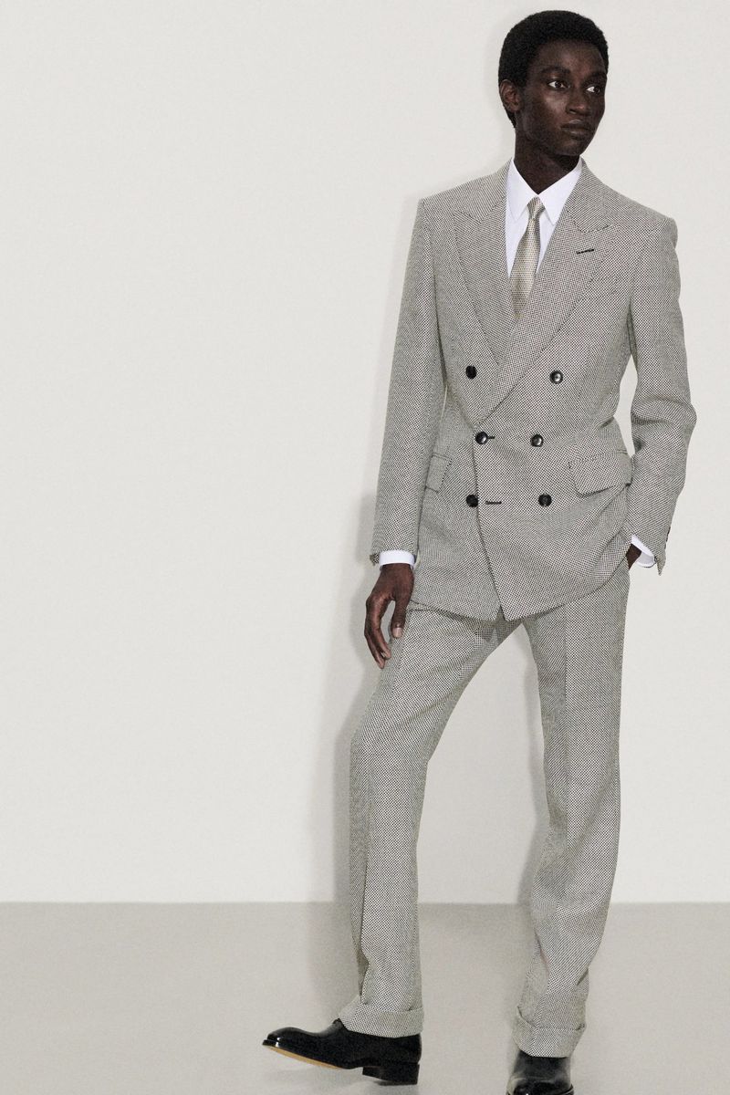 A slender man in a patterned grey double-breasted suit, white shirt, and pale tie stands against a neutral backdrop in a studio shot.