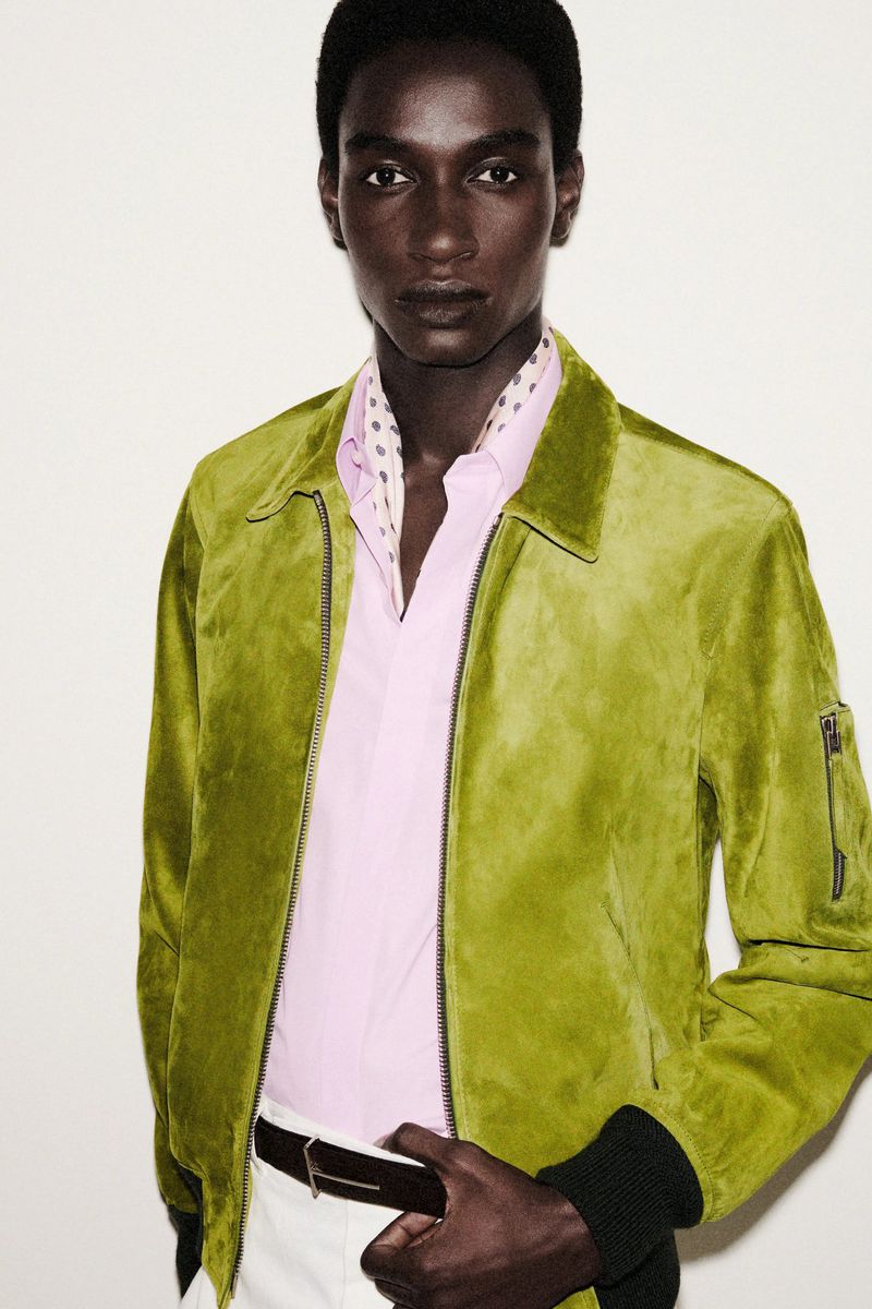 Dark-skinned male fashion model poses indoors wearing a green jacket, light pink shirt, white pants, and a patterned scarf.