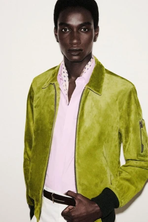 Dark-skinned male fashion model poses indoors wearing a green jacket, light pink shirt, white pants, and a patterned scarf.