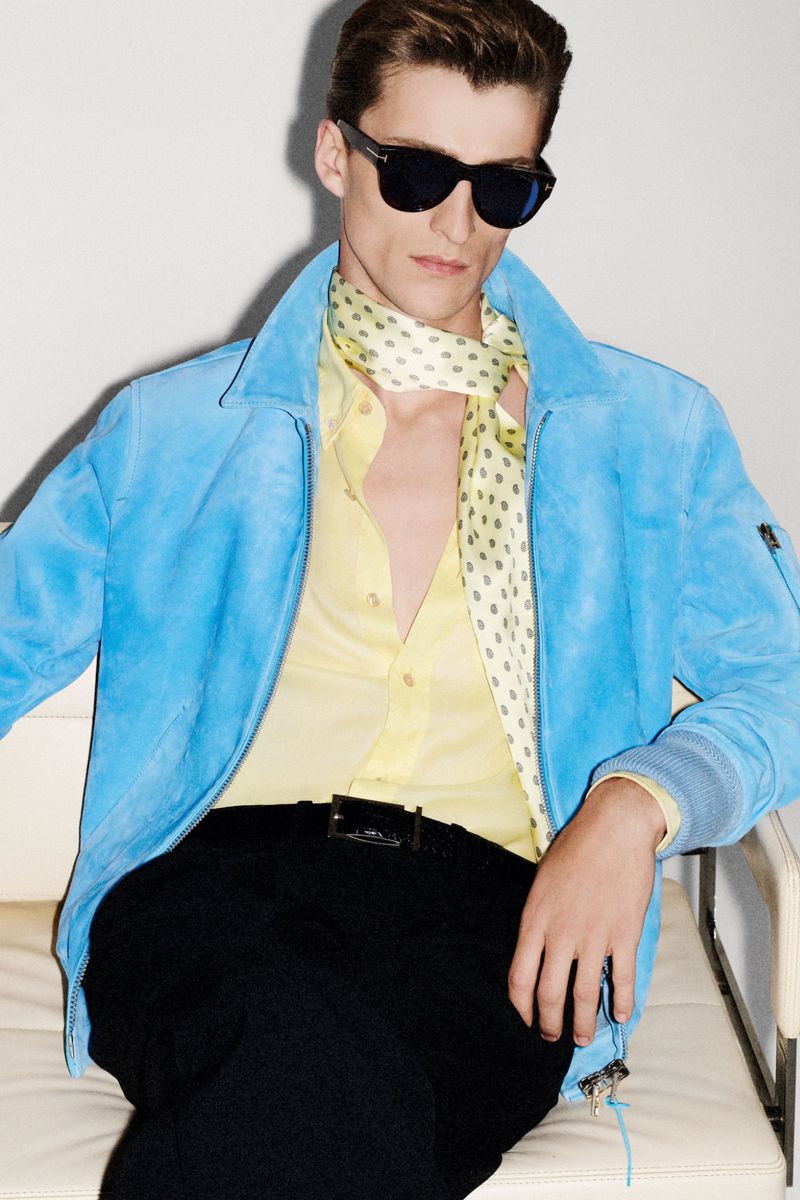 A stylish adult male model sits in a photo studio wearing a blue suede jacket, yellow button-down shirt, black pants, and sunglasses.