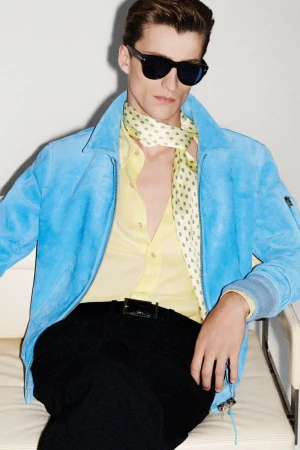 A stylish adult male model sits in a photo studio wearing a blue suede jacket, yellow button-down shirt, black pants, and sunglasses.
