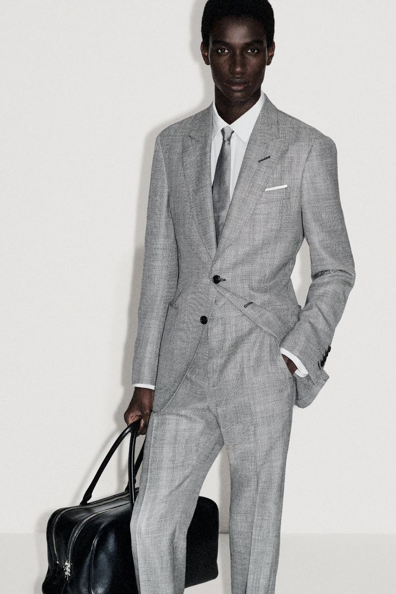 An African-American male model poses in a grey suit with a tie and carries a black leather duffel bag against a plain white background.