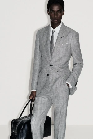 An African-American male model poses in a grey suit with a tie and carries a black leather duffel bag against a plain white background.