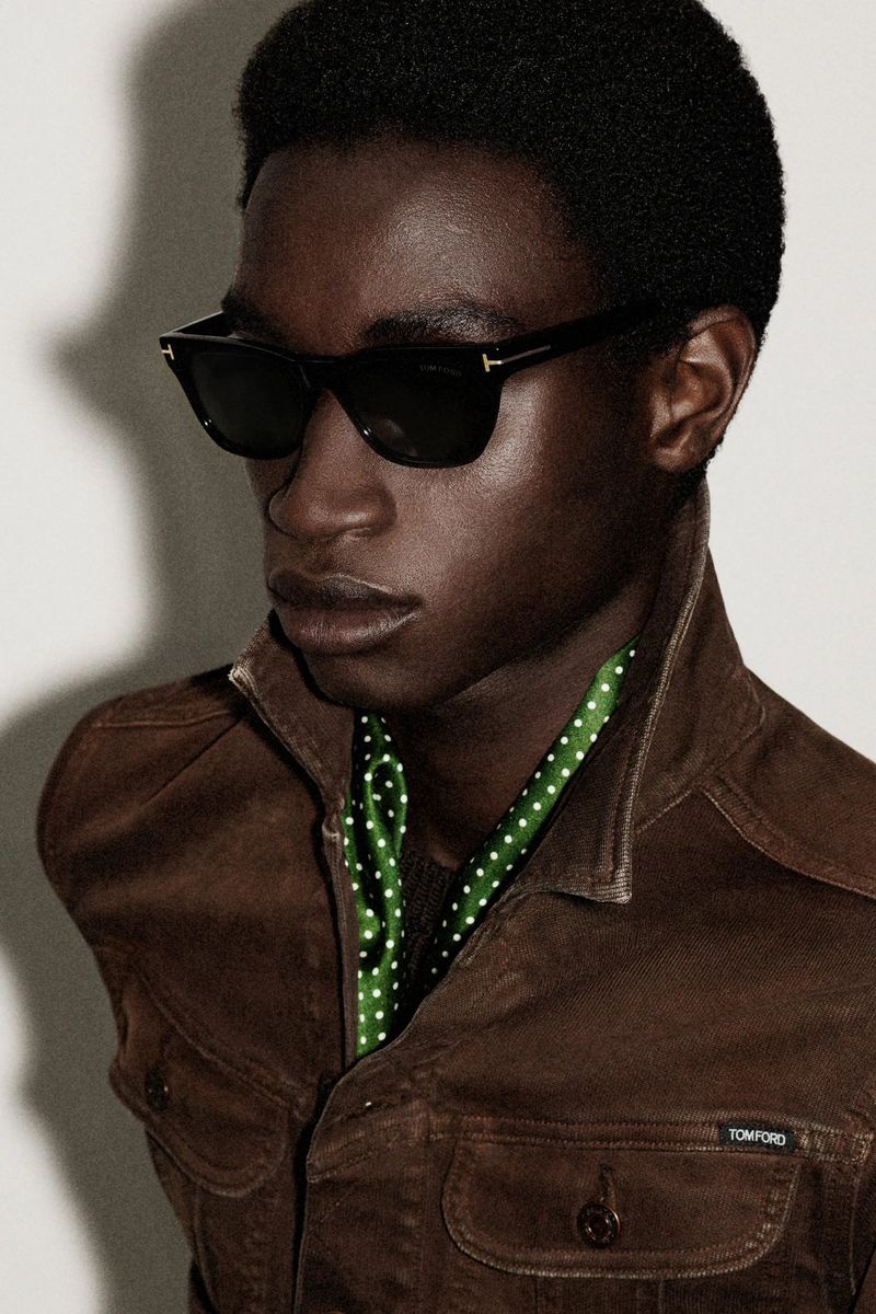 Close-up portrait of a dark-skinned man wearing sunglasses and a brown jacket with a green scarf, showcasing a fashion-forward look.