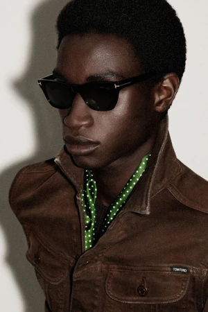 Close-up portrait of a dark-skinned man wearing sunglasses and a brown jacket with a green scarf, showcasing a fashion-forward look.