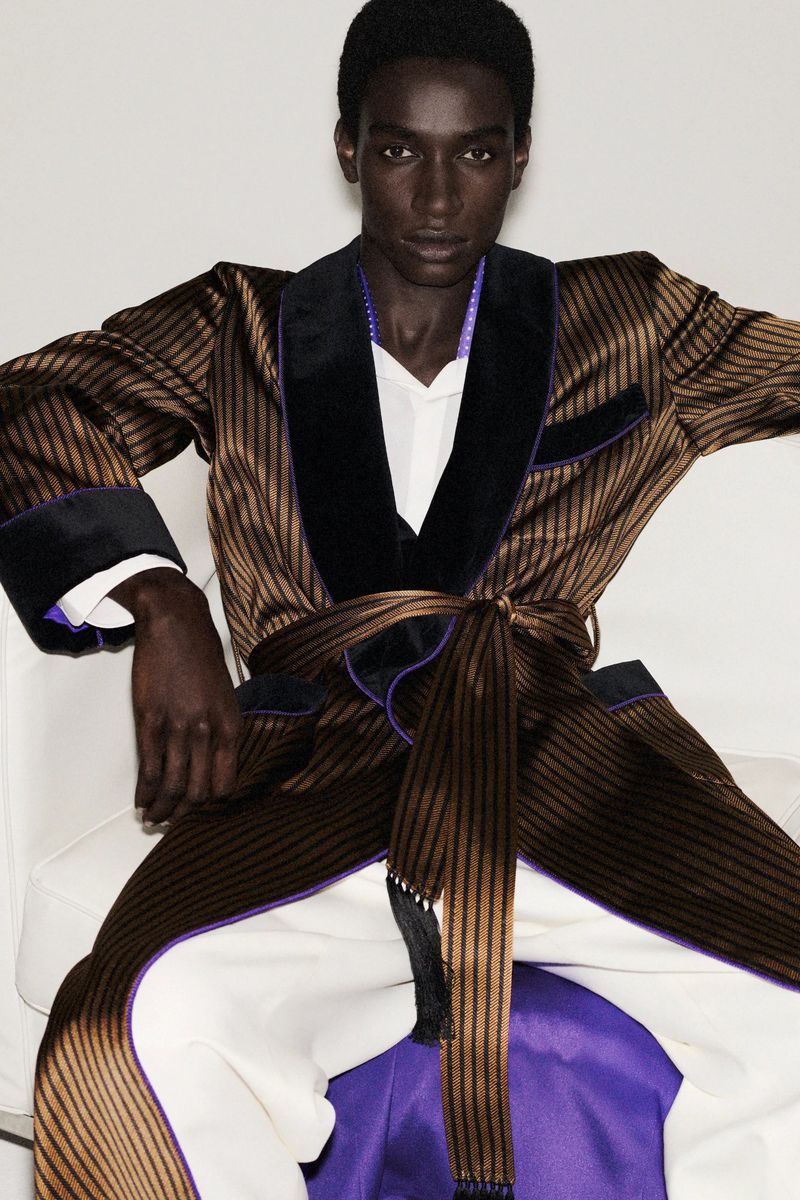 Dark-skinned adult male poses indoors wearing a striped brown robe with black lapels and white pants, seated against a white backdrop.