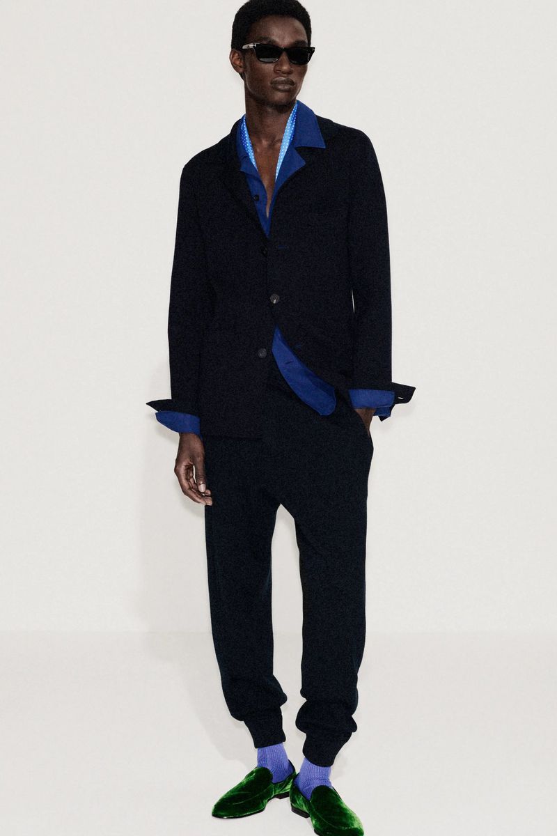 An adult Black male is standing wearing a dark blazer, blue shirt, dark pants, dark sunglasses, and green velvet loafers on a white background.