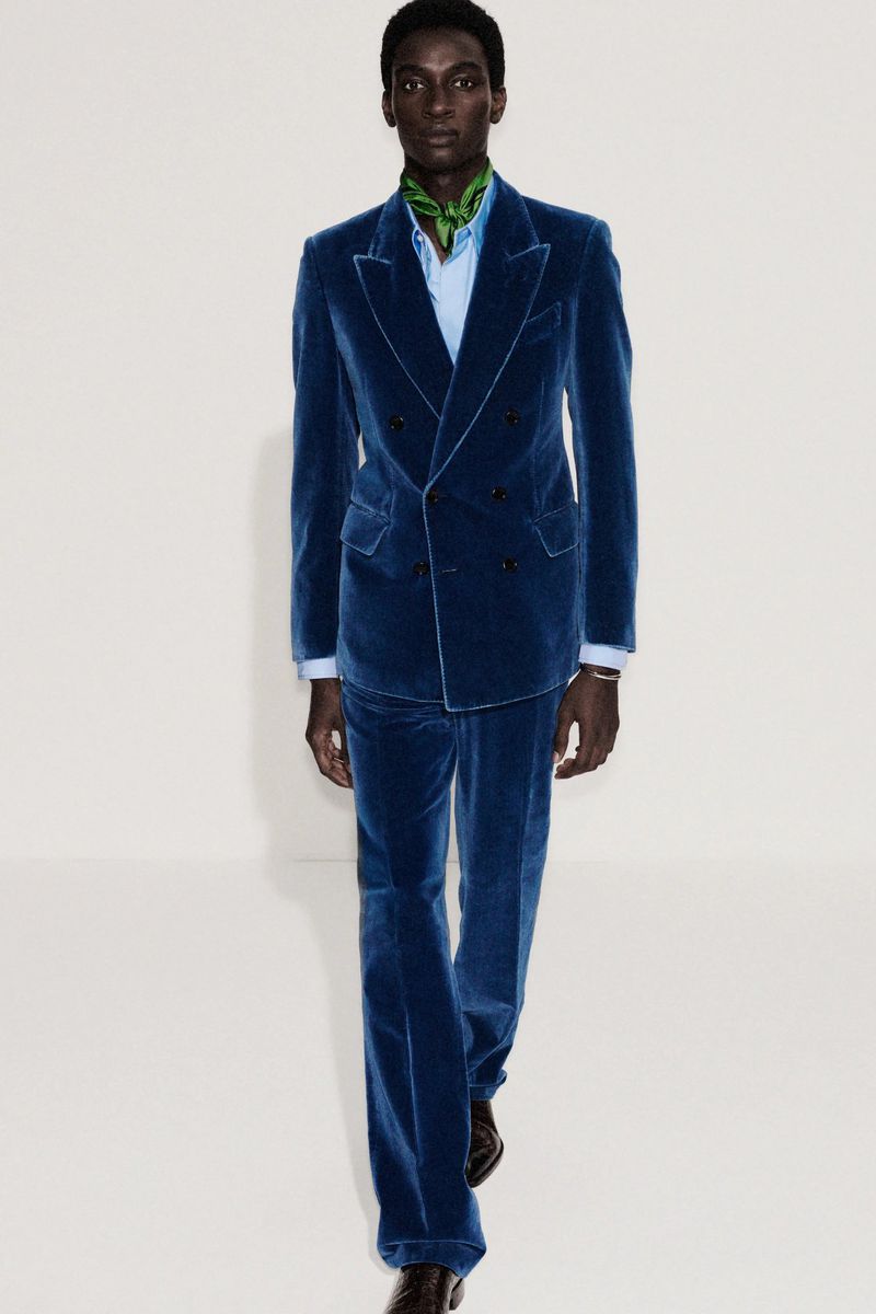 Dark-skinned male fashion model walks in blue velvet suit, light blue shirt, and green scarf, set against a neutral backdrop.