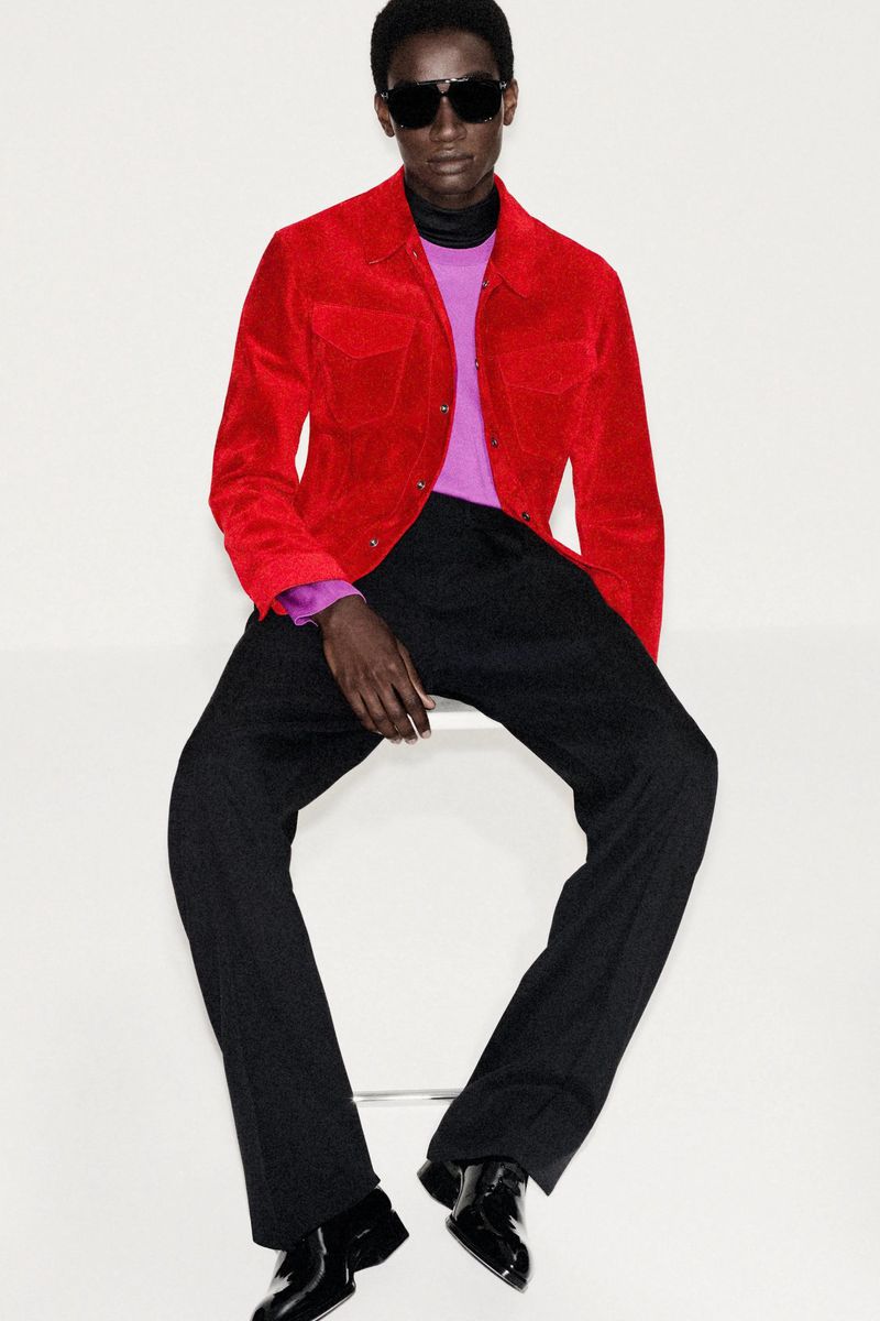 Fashionable Black man poses in a red corduroy jacket, black turtleneck, purple sweater, and black dress pants against a white background.