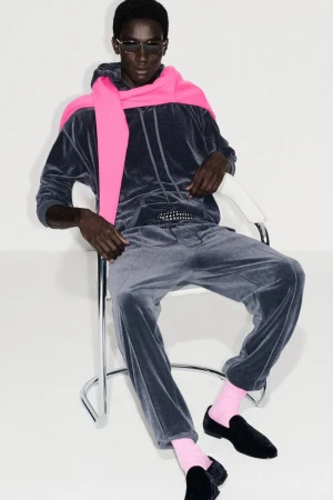 Adult African-American man poses in a chair wearing a velour sweatsuit, pink scarf, sunglasses, pink socks, and black loafers in a studio.