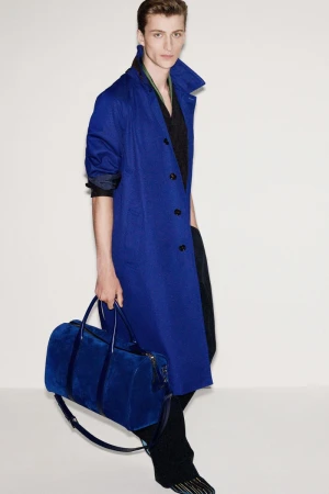 Model in vibrant blue coat carrying matching blue duffel bag against a clean white seamless studio background.