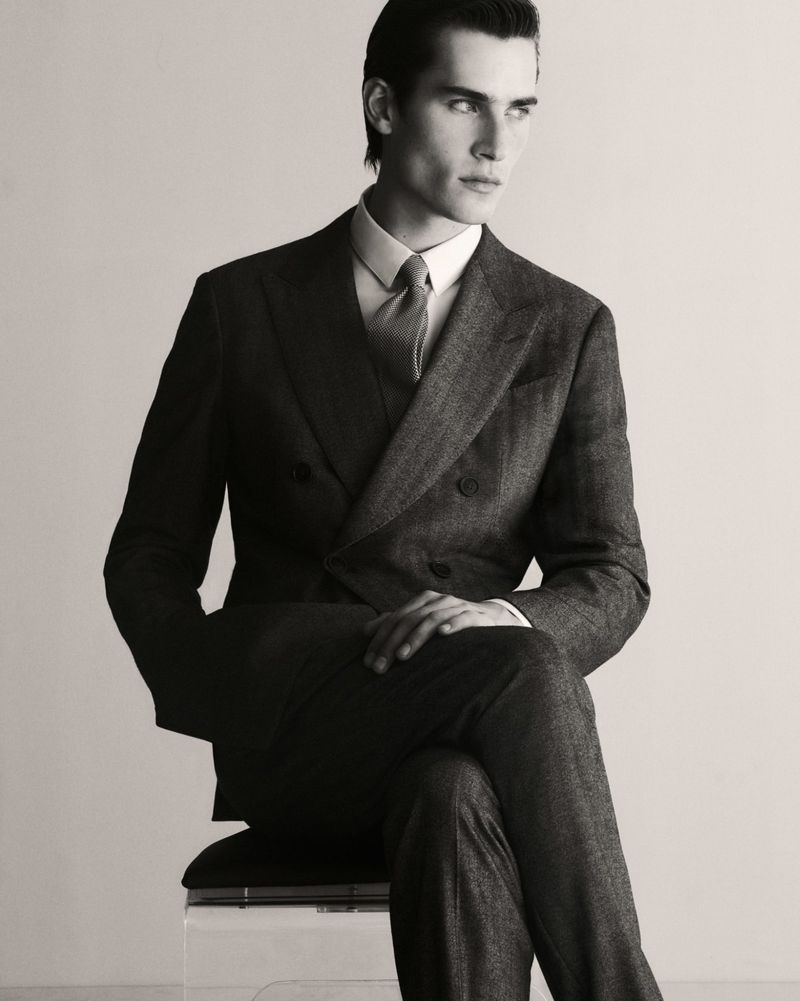 Portrait of an adult male model in a double-breasted suit seated against a white backdrop. The photograph is black and white.
