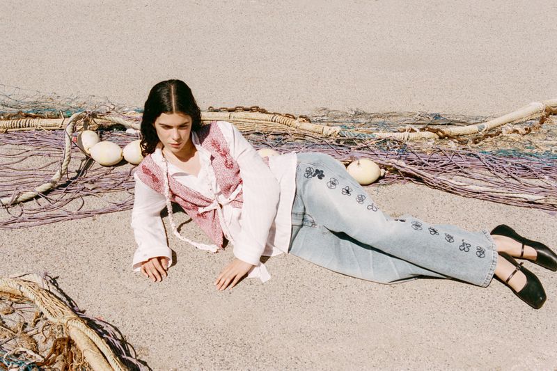 A woman in jeans and a vest lies on a sandy surface near a fishing net and floats, creating a relaxed and artistic fashion shot.