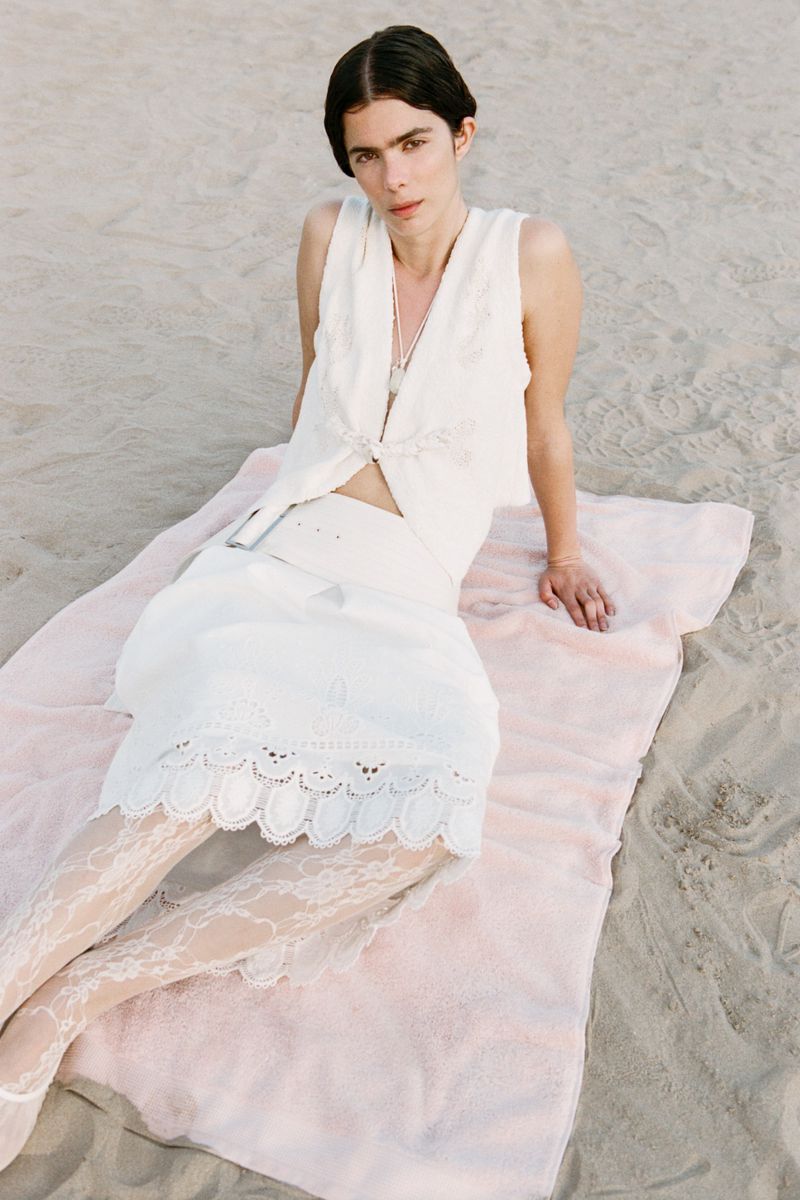 A woman in a white dress and patterned tights sits on a light pink blanket on a sandy beach in an eye-level, medium shot.