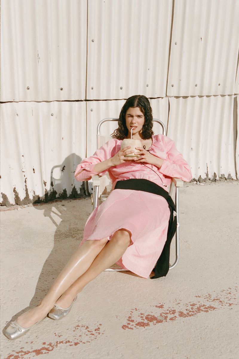 A woman in a pink dress sits in a chair drinking from a coconut with a straw against a textured metal wall outdoors in sunlight.
