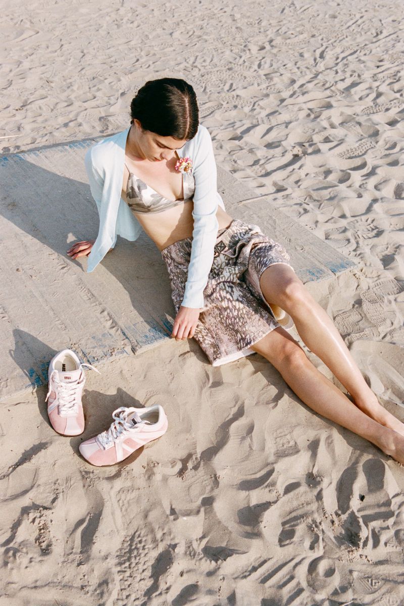 A young woman poses on a beach in a bikini top, skirt, and cardigan, seated on a light gray blanket with pink sneakers nearby.