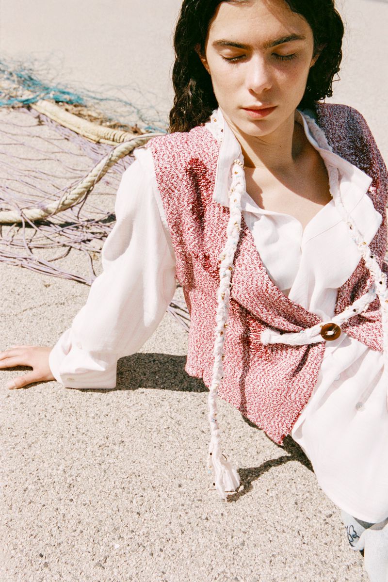 A woman models a sleeveless red and white vest with a white shirt on a beach, seated in the sand with a fishing net in the background.