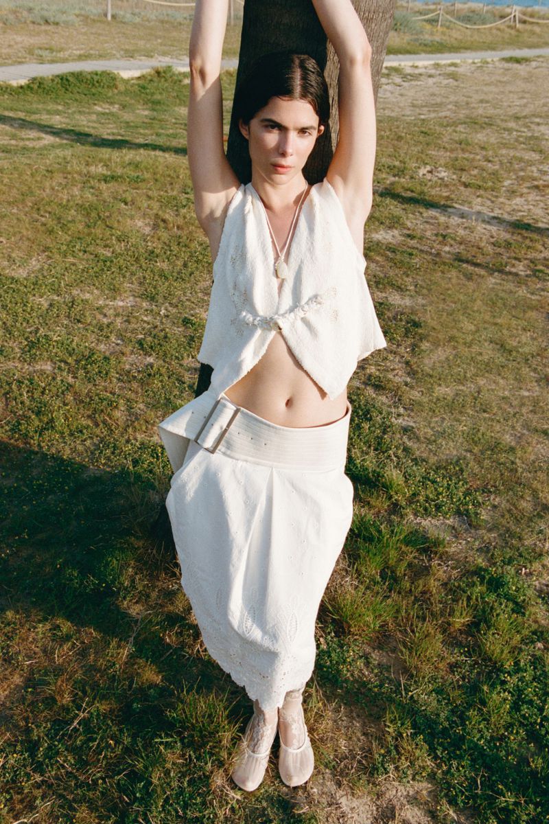 Model Alba Moreira in a white cropped sleeveless vest and matching belted midi skirt posing against a tree outdoors.