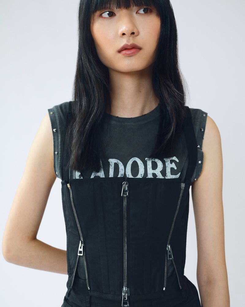 Asian woman in edgy fashion with black zip-front top and studded grey tee posing against a white background.